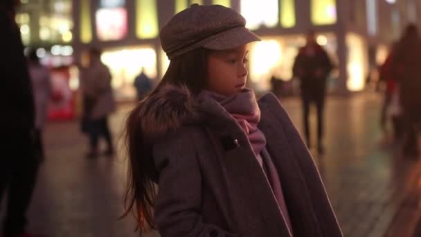 穿着外套 帽子和围巾的女孩站在街上等着 背景是经过人们和傍晚的城市灯光 — 图库视频影像