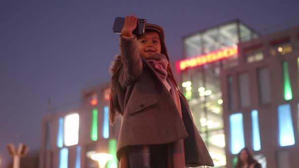 コート マフラーの子女の子のポーズ 笑顔と背景に夜の街の灯の通りで彼女の手で自分のスマート フォンで写真を撮る — ストック動画