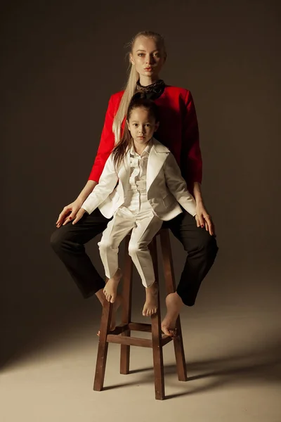 Young mother and little daughter are sitting on chair and posing in studio.