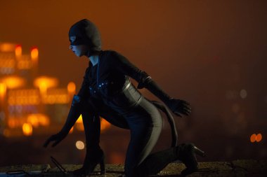 Bir kedi kuyruk, eldiven ve maske, siyah kostümlü kılığında bir kadın gece şehir ışıkları ve gökyüzünün arka planı oturur. Cosplay.