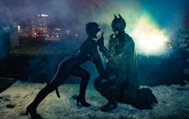 A man dressed as a bat and a woman dressed as a cat stand next to each other on background of night city lights, smoke and buildings. Cosplay. clipart