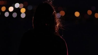 Düşünceli kadın gece gece şehir ışıkları çerçevede rüzgarda duruyor.