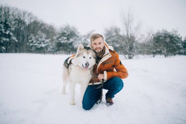 Genç adam bir köpek Alaskan Malamute yanındaki karda oturur ve onun kışın yürüyüşe sarılarak.
