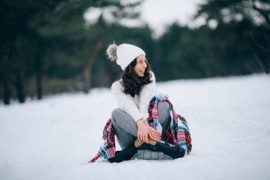 Güzel kadın oturuyor ve kar üzerinde gülüyor beyaz kazak, şapka ve atkı giymiş kış yürüyüş sırasında.
