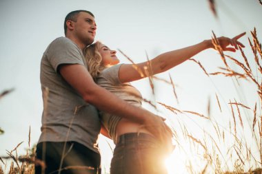 Genç bir adam onun hamile eşi karın arka plan gökyüzü ve spikelets karşı bir yürüyüş sırasında kucaklar. Arka ışık.