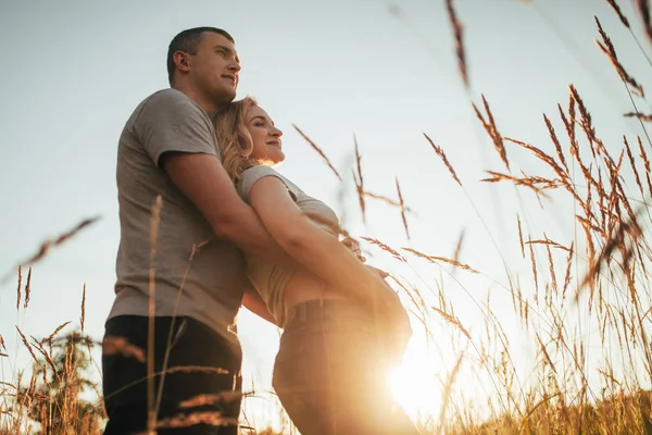 Genç bir adam onun hamile eşi karın arka plan gökyüzü ve spikelets karşı bir yürüyüş sırasında kucaklar. Arka ışık.