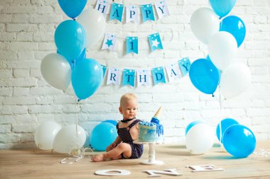 Bir yıl yaşlı erkek çocuk mavi ve beyaz balonlar, Festival pasta ve tuğla duvar ve zemin üzerinde yazıtlar bir sürü arasında ilk Doğum günü kutluyor.