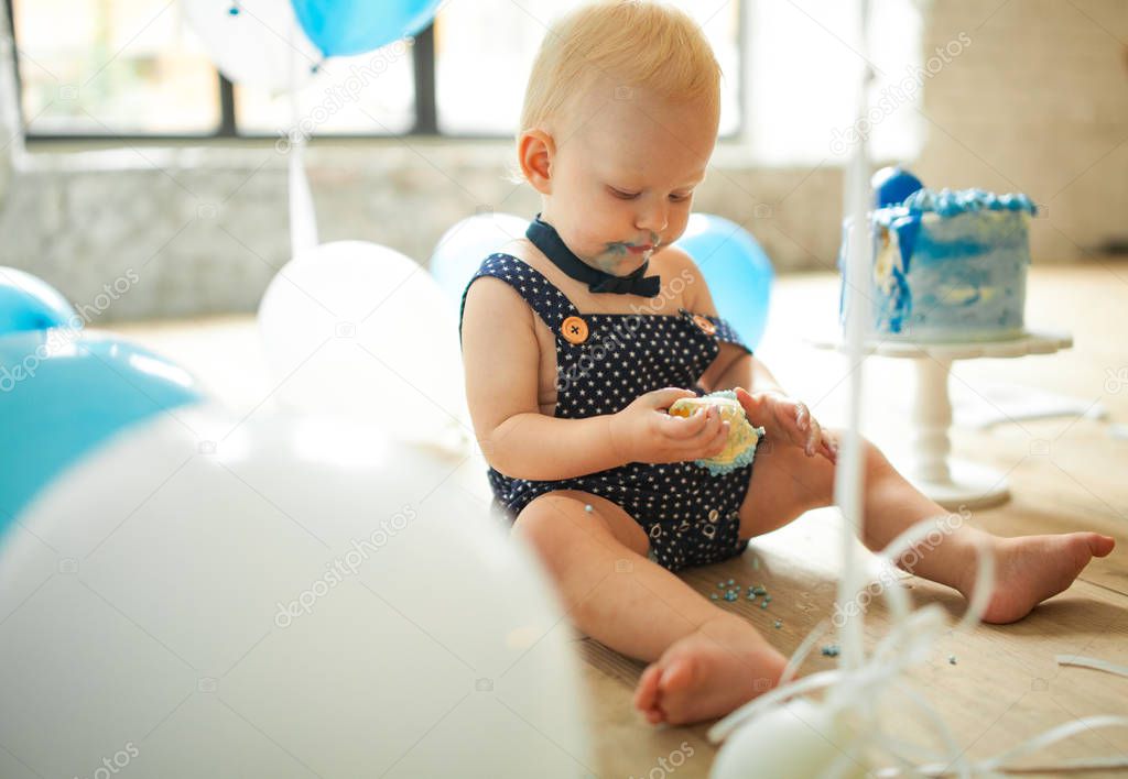 One year old boy is celebrating his first birthday, eating and smashing festive cake on background of blue balloons.