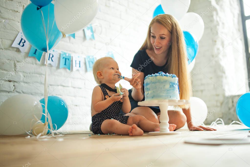Mother is celebrating first birthday of her one year old son and feeding his by festive cake on background of blue balloons.