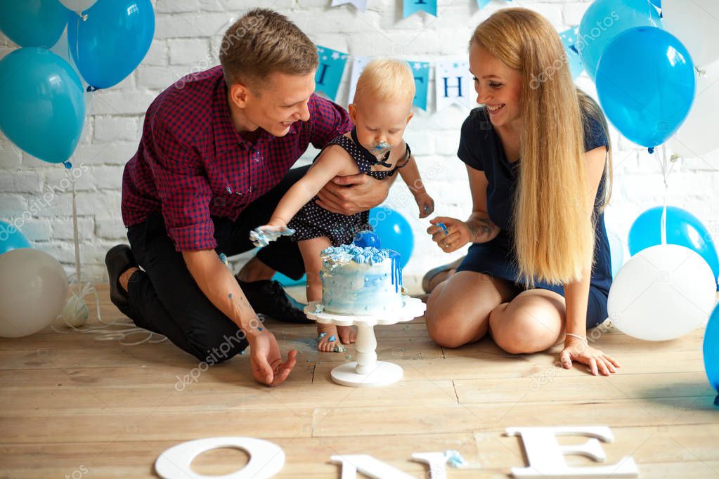 Parents are celebrating first birthday of their one year old son and feeding his by festive cake on background of blue balloons.