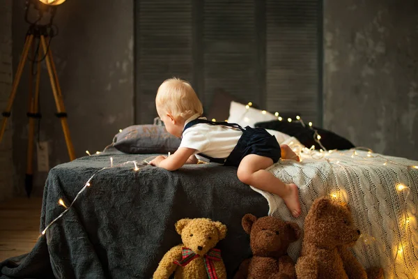 男の子がベッドでクロールし おもちゃの横に光る電球のガーランドで遊ぶ — ストック写真