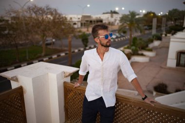 Genç adam beyaz gömlek ve güneş gözlüğü Caddesi'nin arka plan üzerinde balkonunda duruyor.