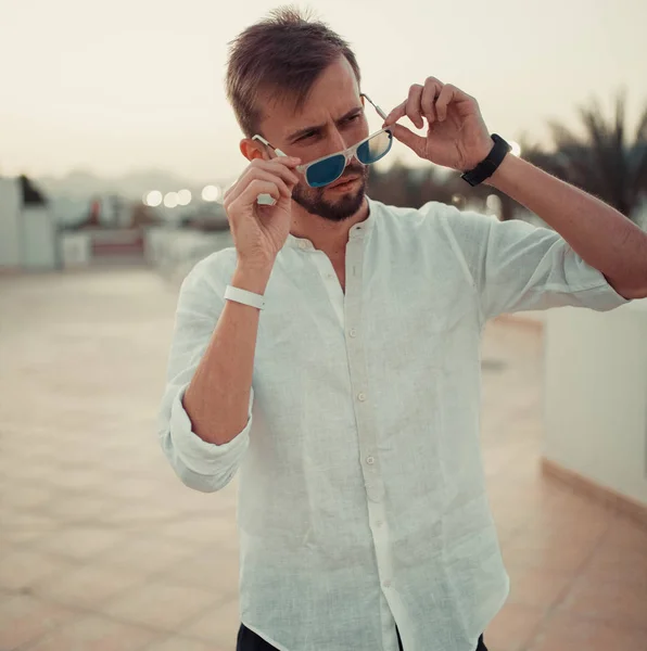 Young man stands on the roof of building in white shirt and sunglasses at sunset.