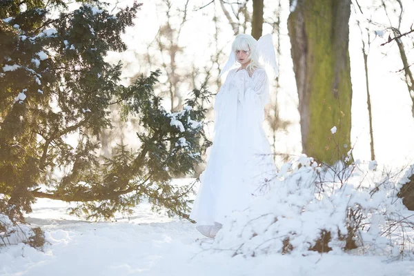 Beautiful blonde girl stands in snowy forest in image of good angel with wings dressed in white clothing.