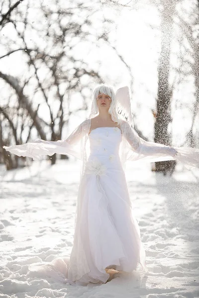 Beautiful blonde girl stands in snowy forest in image of good angel with wings dressed in white clothing against background of snowfall.