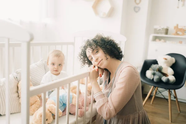 Lycklig mamma sitter bredvid sitt barn i spjälsängen. — Stockfoto