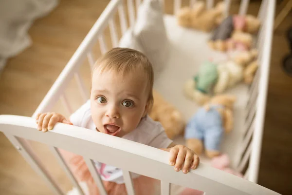 Niña pequeña se para en la cuna entre los juguetes . —  Fotos de Stock