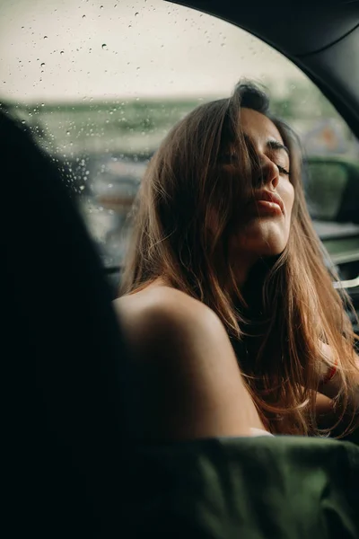 Uma jovem se senta dentro de um carro contra o pano de fundo de gotas de chuva — Fotografia de Stock