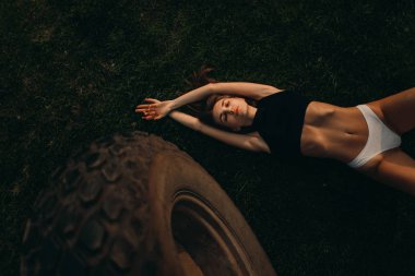 Bir kadın büyük bir tarım makinesi çarkının yanında çimlerin üzerinde yatıyor.