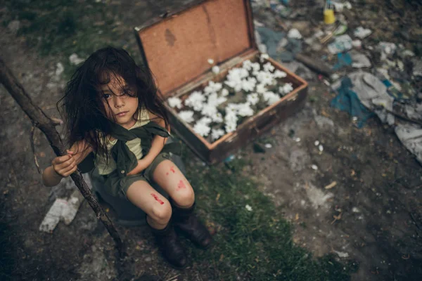 Una chica sin hogar está sentada en un basurero junto a una maleta con flores dentro. . — Foto de Stock