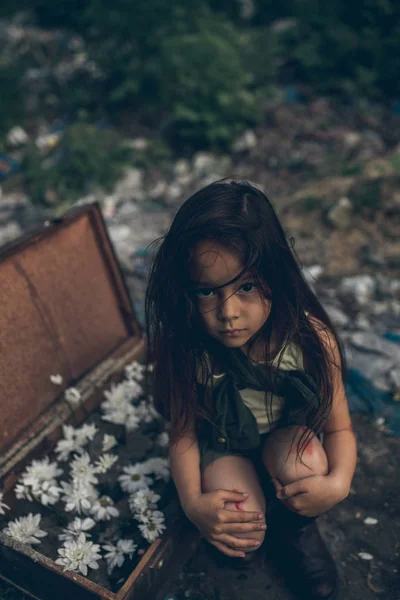 Una chica sin hogar está sentada en un basurero junto a una maleta con flores dentro. . — Foto de Stock