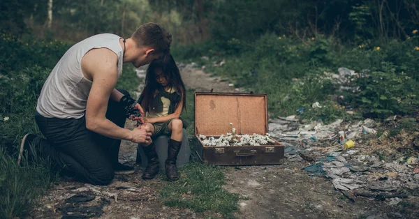 Homeless father and daughter are in garbage dump next to a suitcase with flowers inside. — Stock Photo, Image