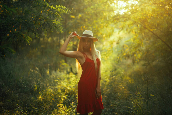 A young woman in a red dress and hat is standing in the forest lit by the sun.