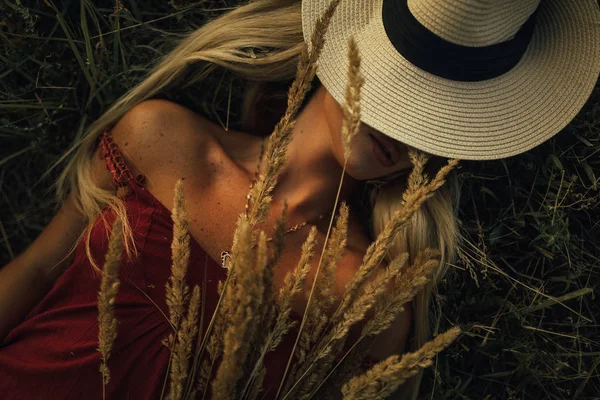 Ung kvinna i en hatt ligger på ängen bland gräs öronen. — Stockfoto
