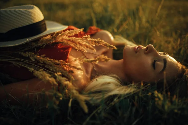 Νέα ξανθιά γυναίκα με καπέλο είναι ξαπλωμένη στο λιβάδι. — Φωτογραφία Αρχείου