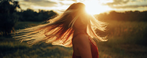 Young blond woman stands on meadow with loose hair lit by sun at sunset. Backlit. Closeup.