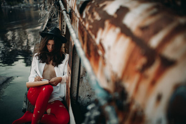 A young woman sits and draws in sketchbook on an old abandoned ship in an unbuttoned white shirt, hat and red holey tights.