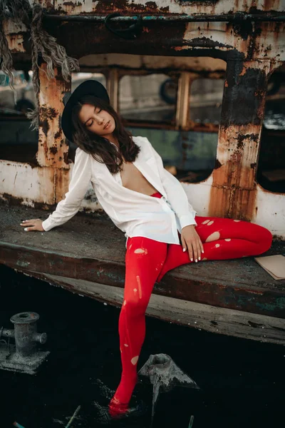 Mladá žena sedí na staré opuštěné lodi v košili, klobouku a červených punčoštech.. — Stock fotografie