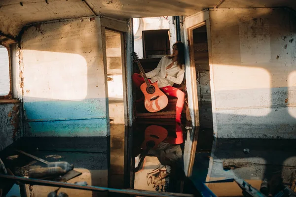 Genç bir üzgün kadın eski terk edilmiş geminin içinde gitar ile oturur. — Stok fotoğraf