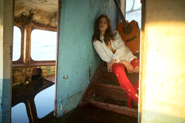 Mladá smutná žena sedí s kytarou uvnitř staré opuštěné lodi. — Stock fotografie