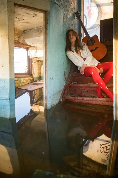 Mladá smutná žena sedí s kytarou uvnitř staré opuštěné lodi. — Stock fotografie