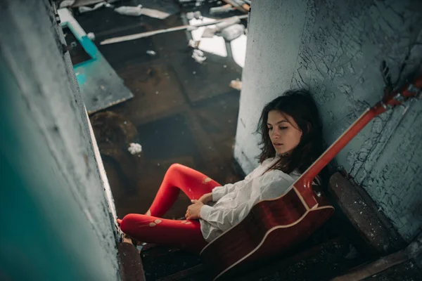 Mladá žena sedí s kytarou uvnitř staré opuštěné lodi. — Stock fotografie
