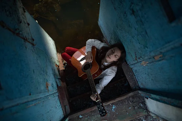 Mladá žena sedí a hraje na kytaru ve staré opuštěné lodi. — Stock fotografie