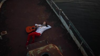 Genç bir kadın su arka plan karşı beyaz gömlek ve kırmızı delikli tayt eski bir terk edilmiş gemide gitar ile yatıyor. Üstte görüntü.