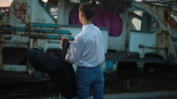 穿白衬衫和破牛仔裤的女孩站在旧废弃的船旁边 把一个装有吉他的箱子放在背上 然后离开 — 图库视频影像