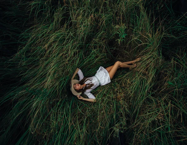 Pretty young woman in hat is lying among the grass on meadow. — Stock Photo, Image