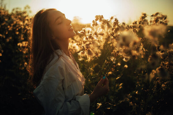 Happy young woman in white shirt is walking with pleasure among the flowers and grass on meadow at sunset.