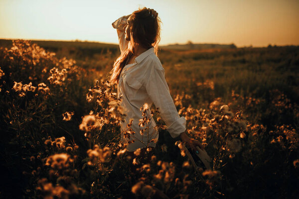 Happy young woman in white shirt is walking with pleasure among the flowers and grass on meadow at sunset.