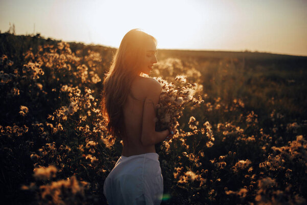 Happy young woman is walking with bouquet in her hands among the flowers and grass on meadow at sunset.