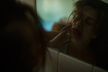 Dayak yiyen kanlı şiddet mağduru kadın aynada yüz yaralarını inceliyor. 