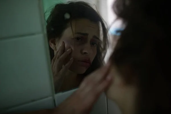 Beaten bloodied woman victim of violence examines her facial wounds in the mirror. — Stock Photo, Image