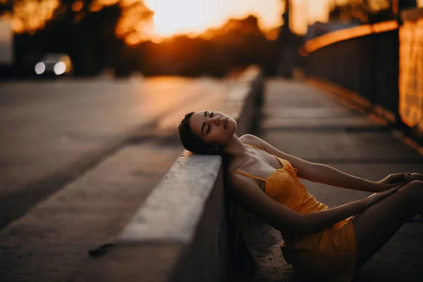 A woman sits on the sidewalk in a yellow dress at sunset. — ストック写真