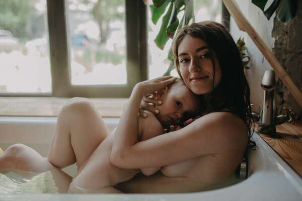 Mother bathes in bathtub and hugs her baby on background of houseplant.