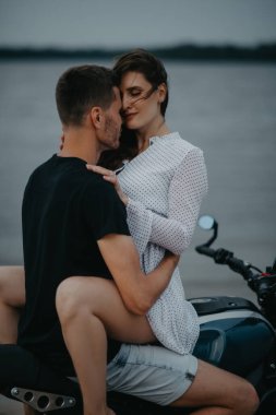 Sahilde iki aşık kucaklaşır, motorsikletin üstünde su üstünde otururlar. Yakın çekim..