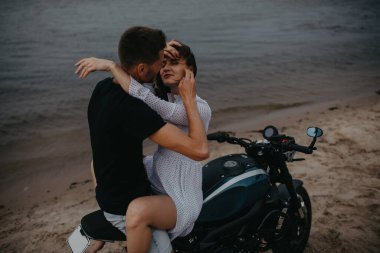 Sahilde iki aşık kucaklaşır, motosiklette su üstünde otururlar..