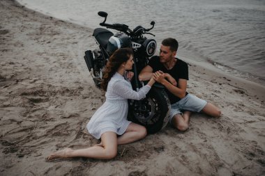 Birbirine aşık çiftler, motorsikletin yanındaki kumsalda oturup el ele tutuşurlar. Üst görünüm.